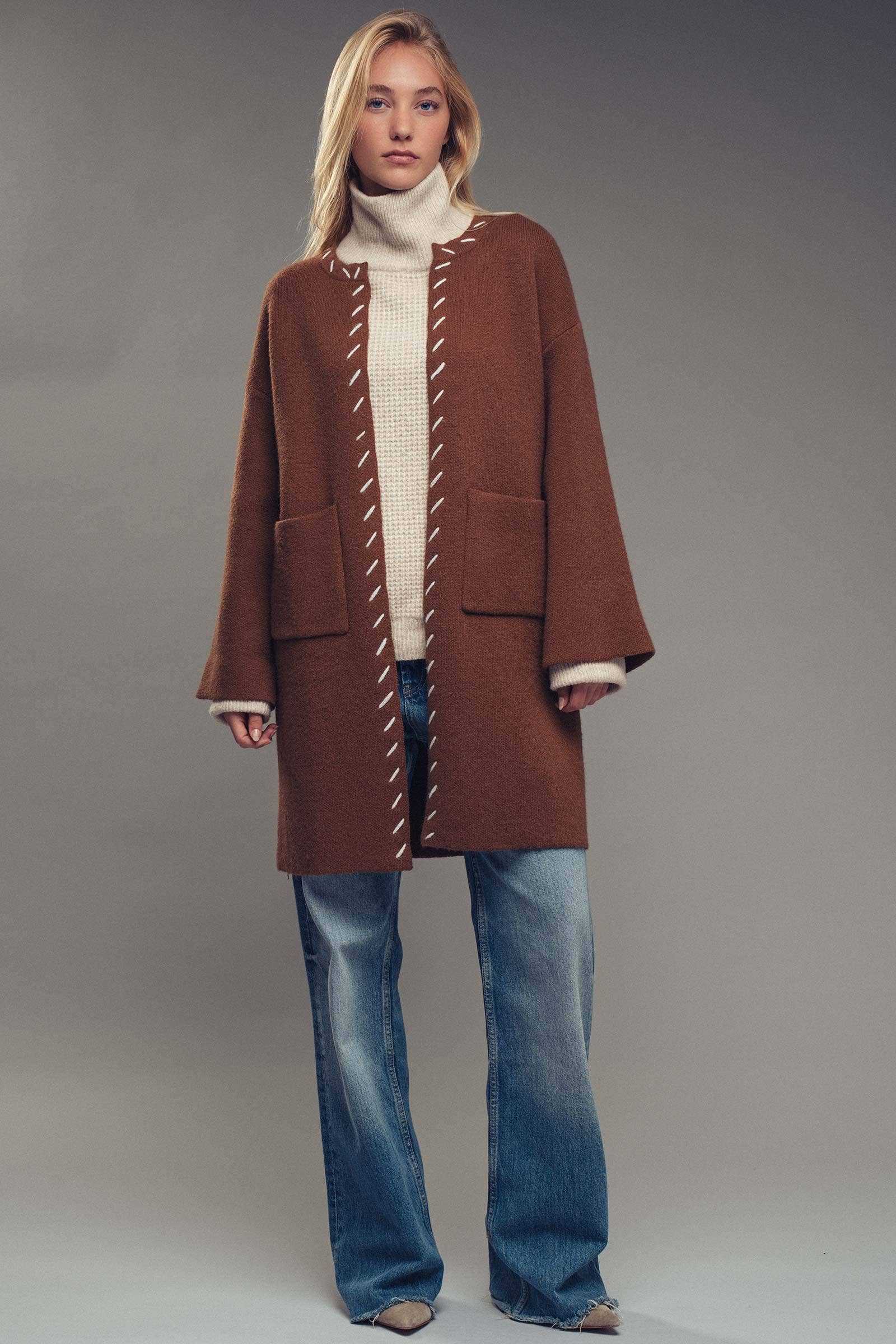 Woman wearing a brown coat with white stitching, beige turtleneck, and blue jeans on a gray background