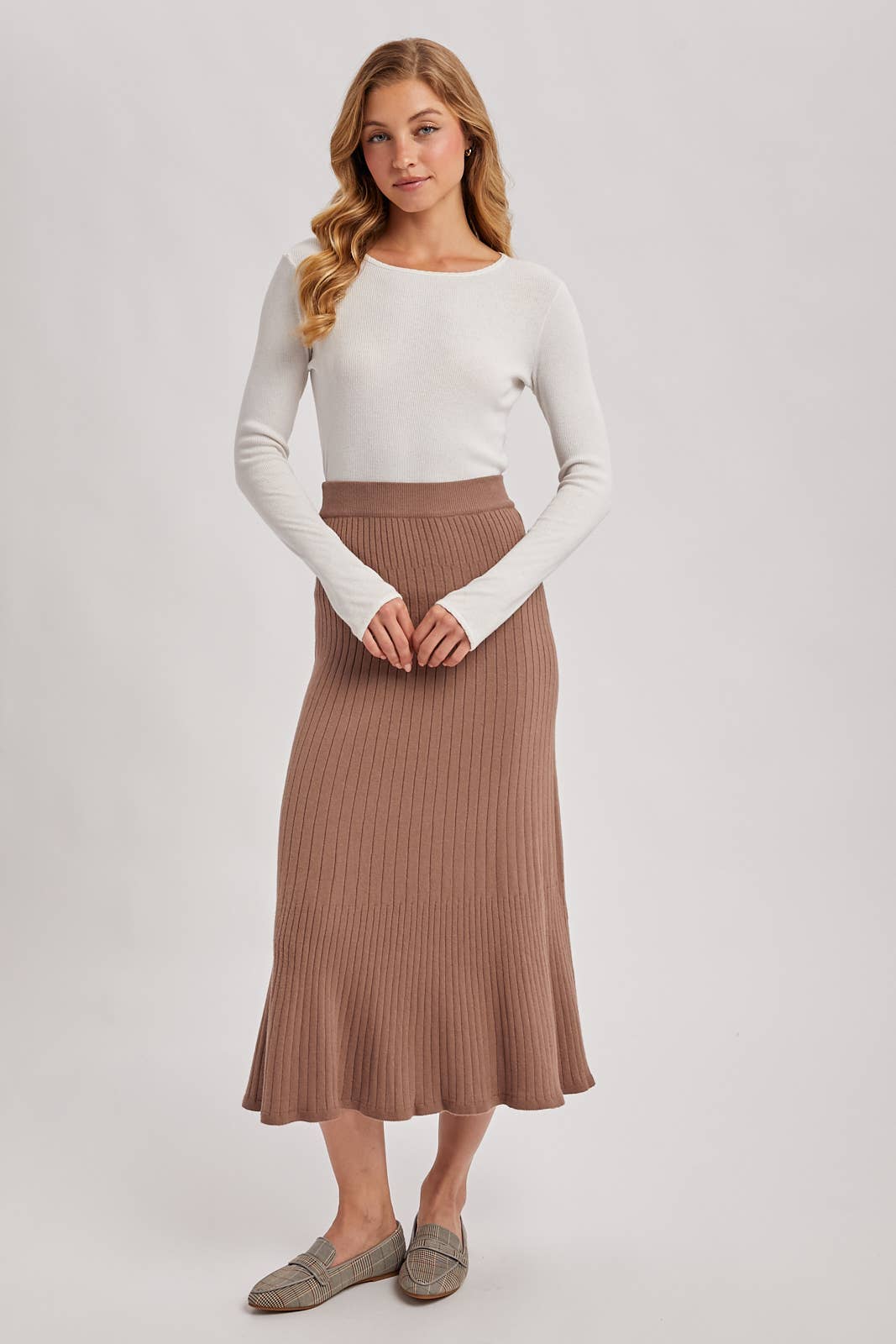 Woman wearing a white long-sleeve top and brown ribbed skirt on a plain background