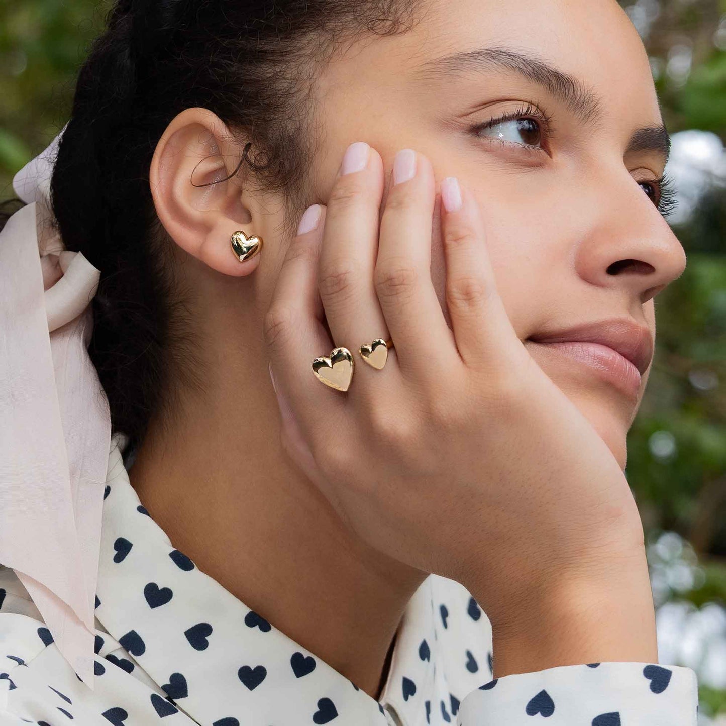 Woman wearing gold heart-shaped earrings and a ring, with a blurred green background