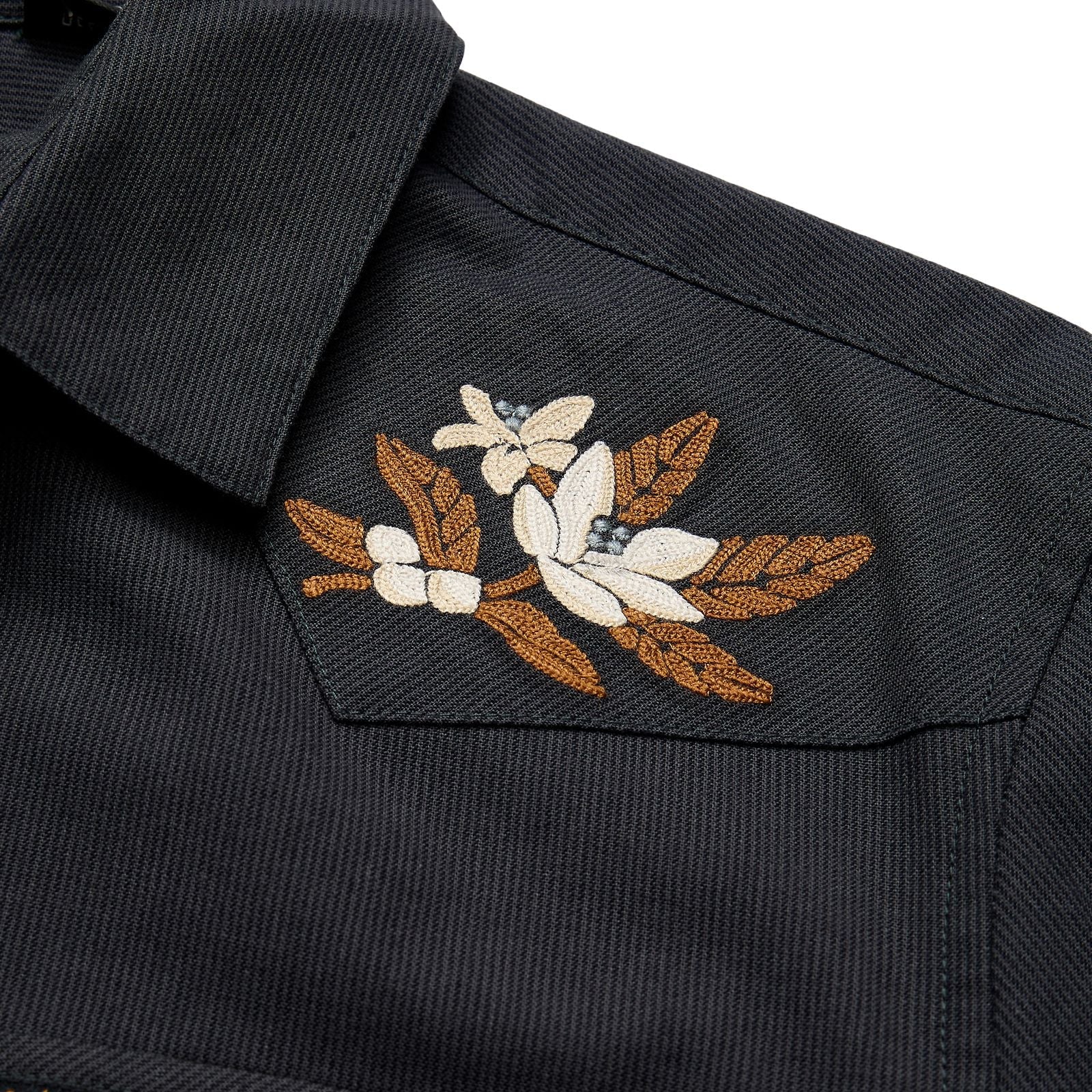 Close-up of a dark charcoal gray textured shirt jacket featuring a detailed floral embroidery on the chest pocket with white flowers, brown leaves, and small blue accents.