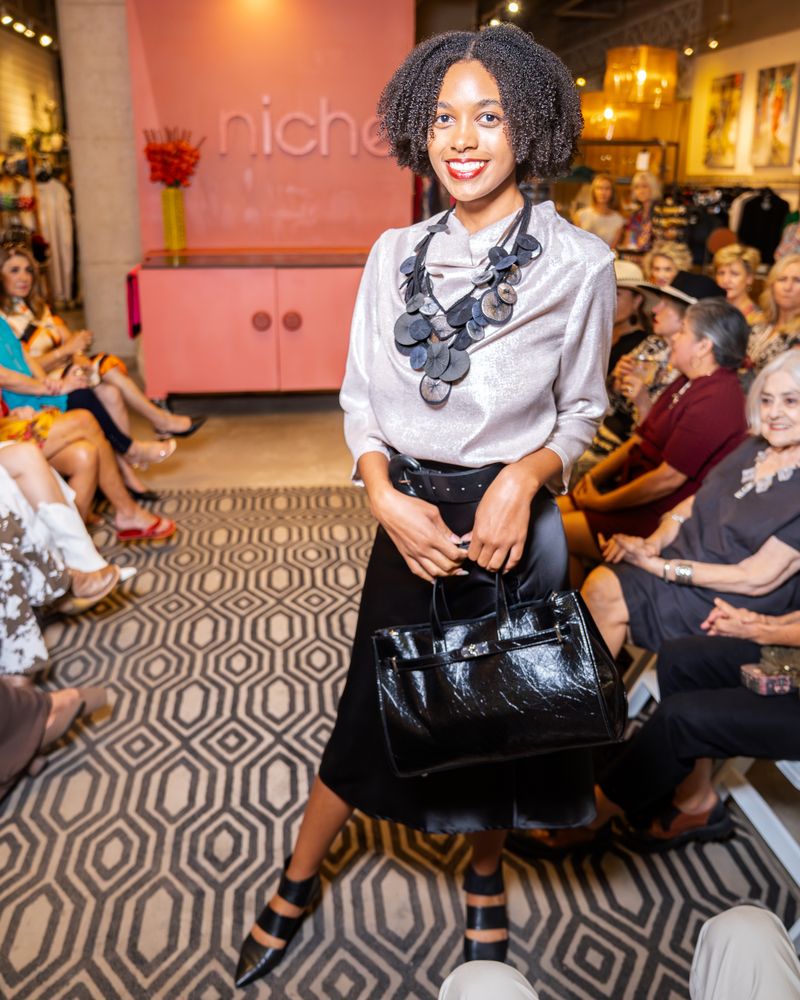 model wearing a silver shimmery blouse, platillos necklace, belt, purse and black satin skirt walking down a runway