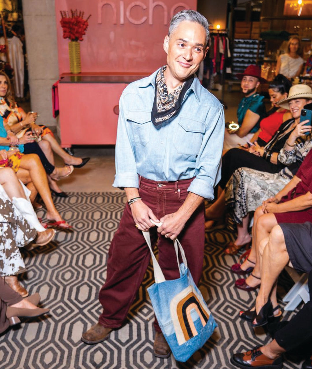 model wearing light blue pearl snap shirt with bandana, burgundy pants and jean tote with rainbow design on a runway