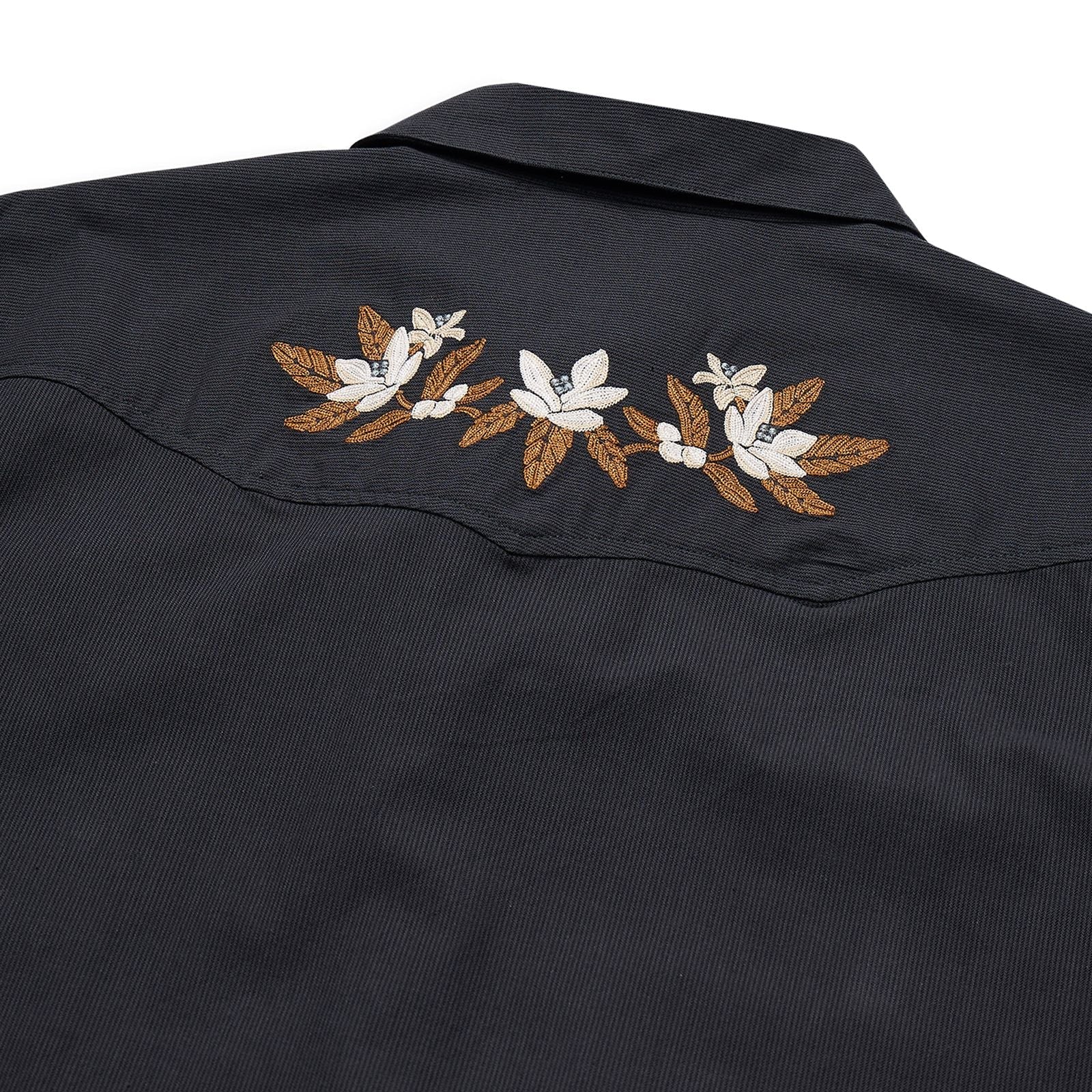 Close-up of a black western-style shirt jacket showing embroidered white flowers and brown leaves across the shoulder yoke, on a white background.