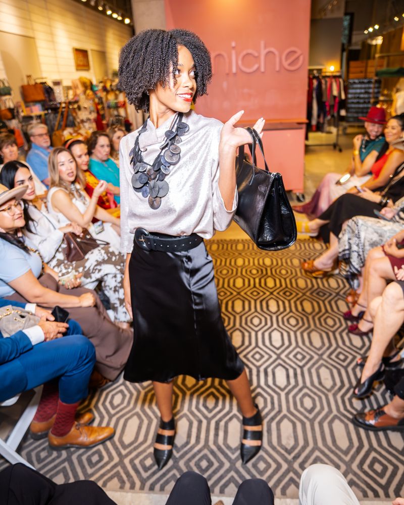 model wearing a silver shimmery blouse, platillos necklace, belt, purse and black satin skirt walking down a runway