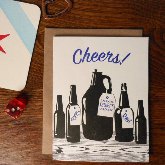 card with bottles on it that says "Cheers!" and "Happy Father's Day" with wood background, dice, coaster, and bottle opener next to it.