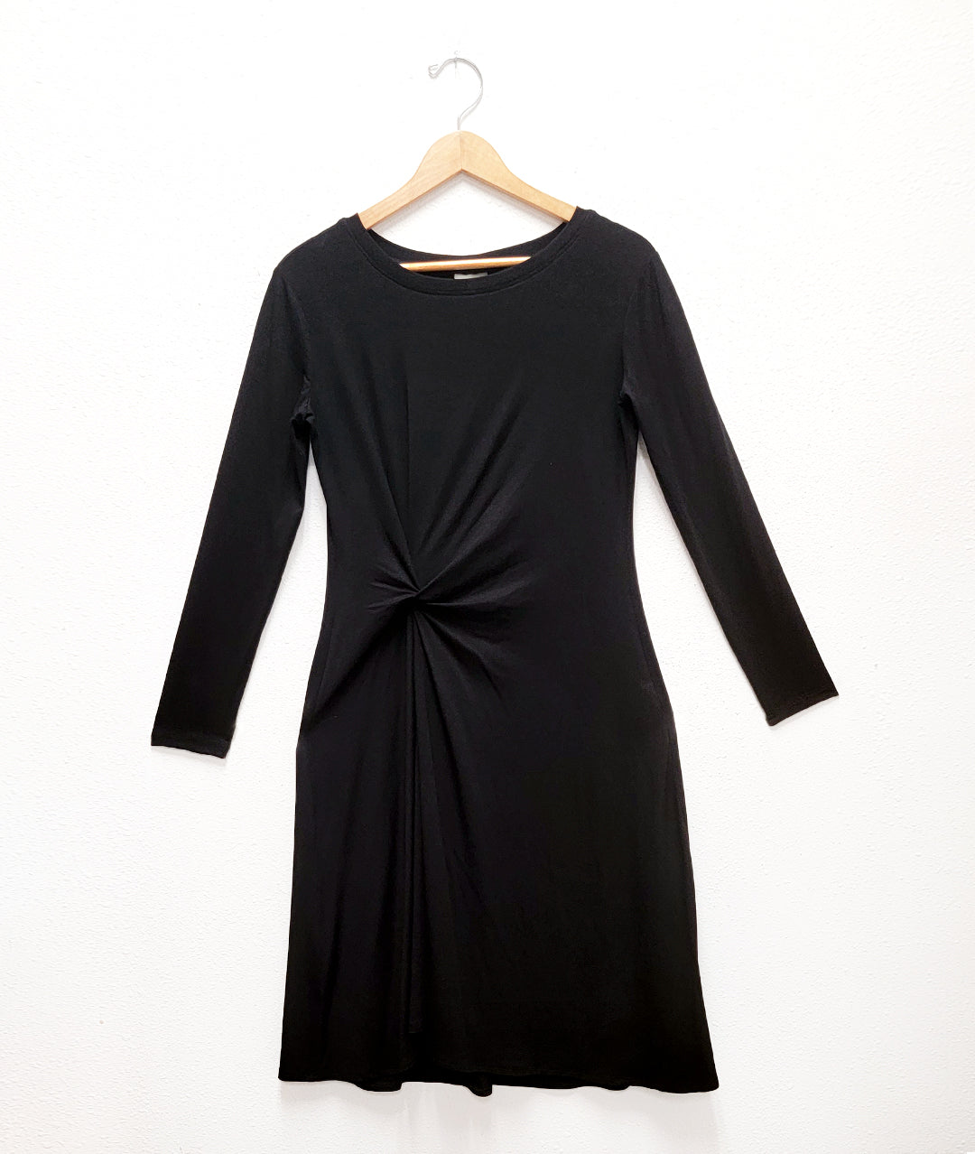 black dress with long sleeves and a twist detail at the waist, on a hanger against a white wall