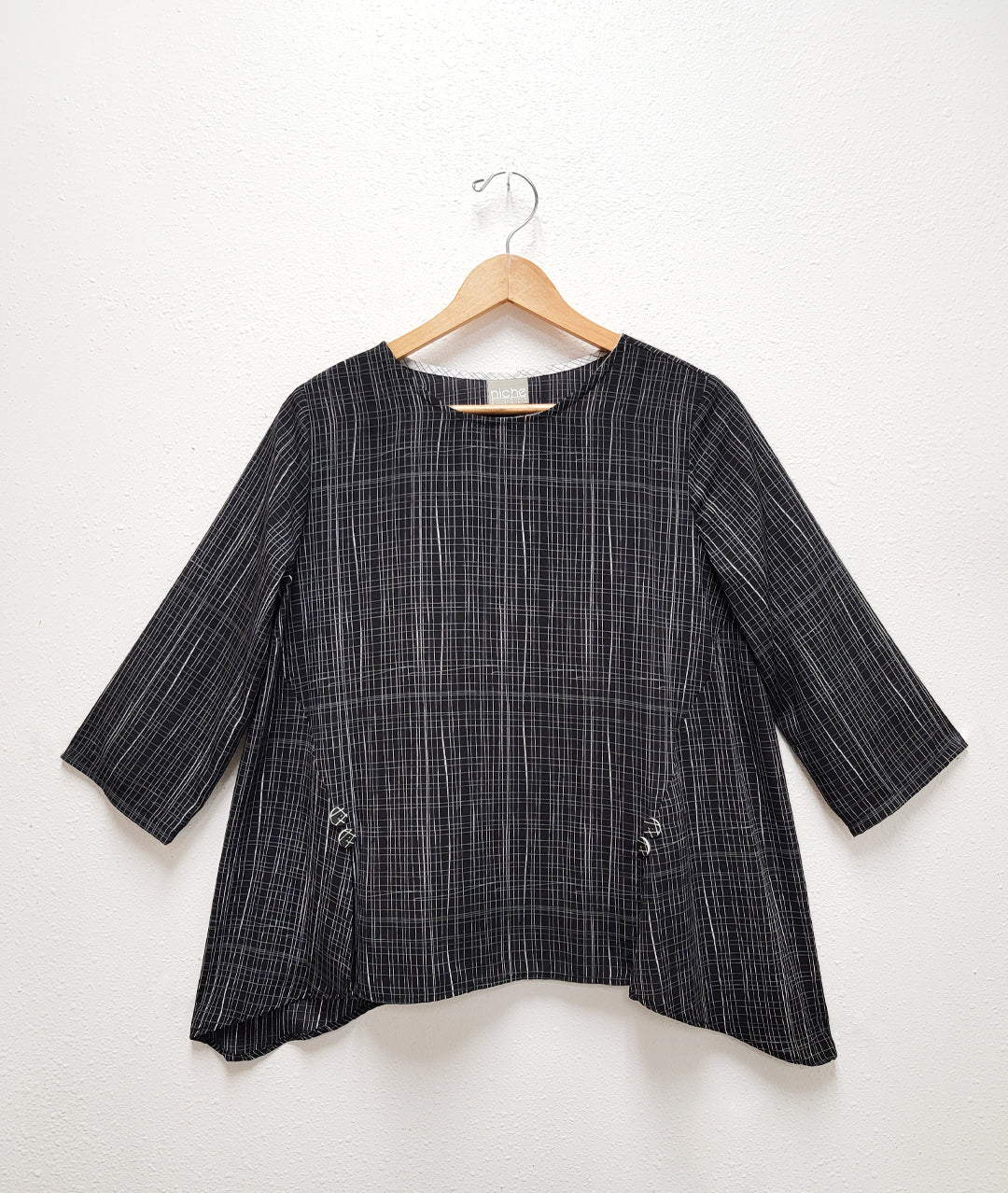 a black pullover top with a white grid print. top has 3/4 sleeves, godets extending from the sides towards the center front, with a split at each seam topped with a button detail