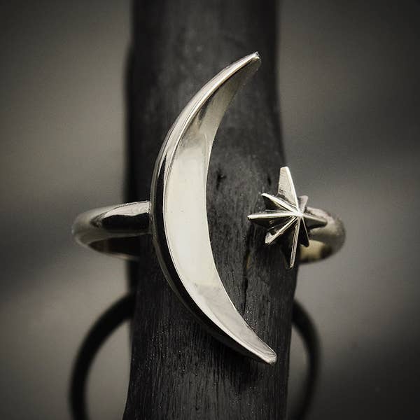 close up of open adjustable ring, one side has a long curved moon shape, other side has 6 point starburst shape. Ring is shown on a dark piece of wood against a shadowy grey background
