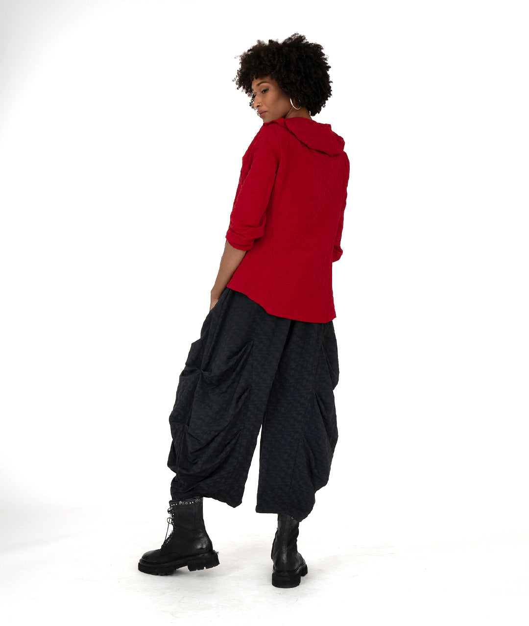 model in a wide leg black pant with a tucking detail along the legs, worn with a red jacket with a ruffled collar and a single button at the neckline
