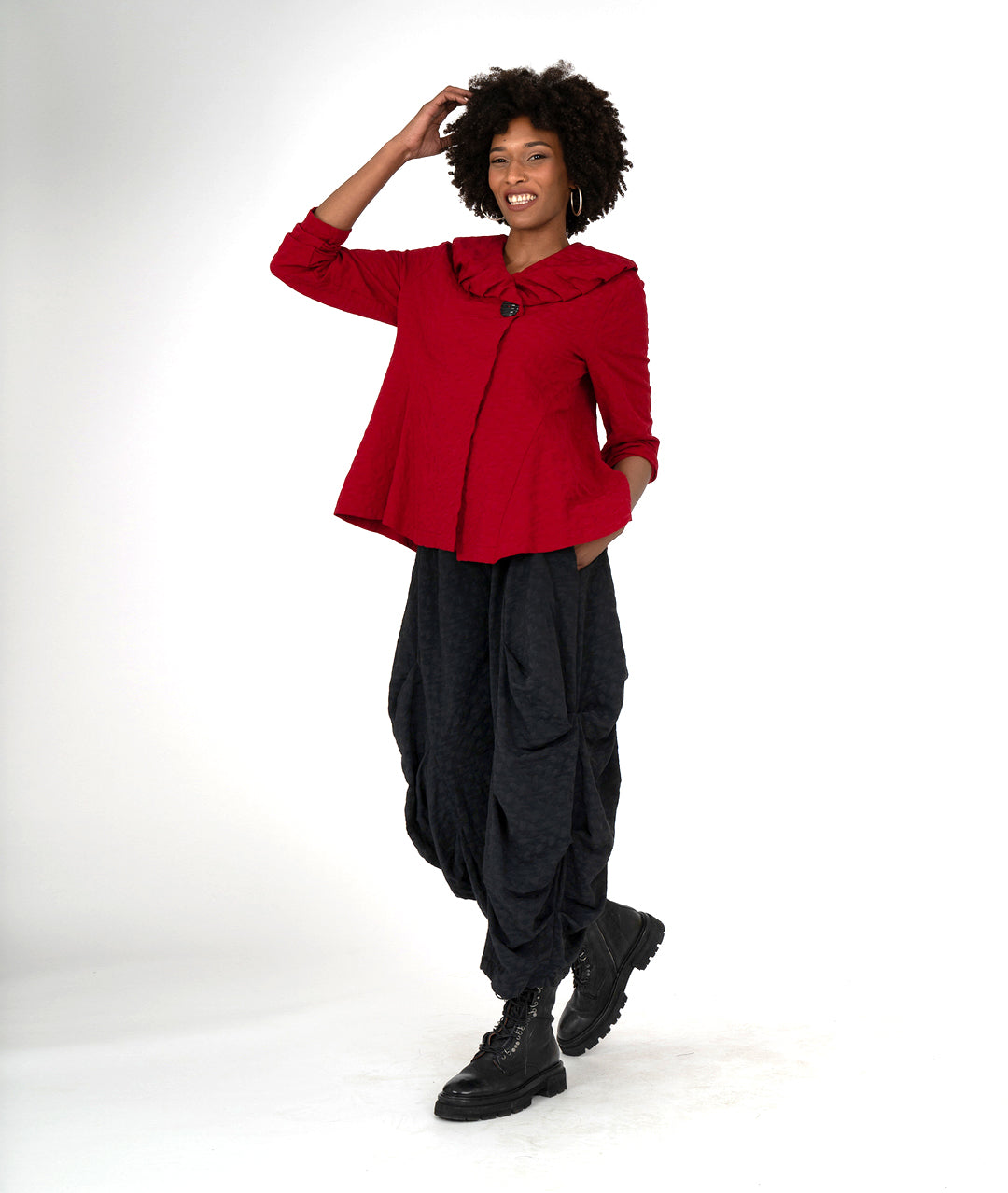 model in a wide leg black pant with a tucking detail along the legs, worn with a red jacket with a ruffled collar and a single button at the neckline
