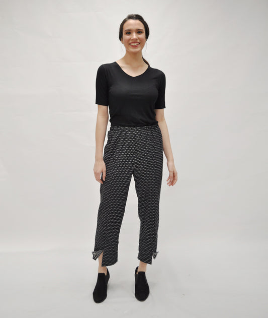 model in a black top with a black pant with small white x's all over. pant has a center front seam with a split at either pant hem, held open with a small button