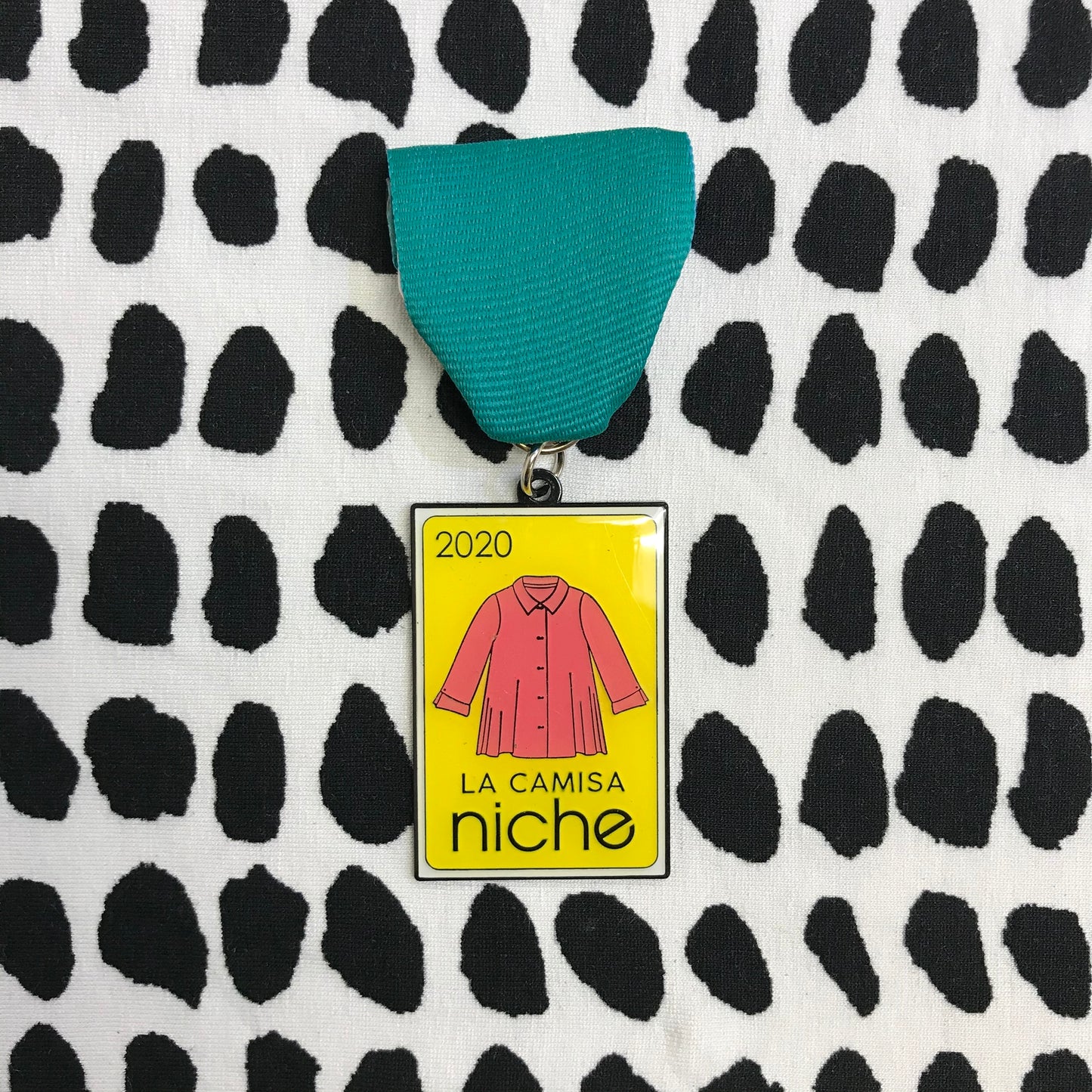 fiesta medal with a teal ribbon on a black and white dot background. The medal is yellow with a coral color blouse and the text "2020" "la camisa" and "niche"