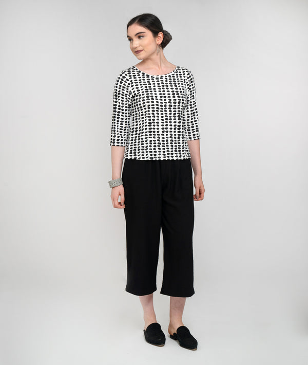 Pictured against a white background is a light skinned model with dark hair. She is looking to her right side, smiling and wearing a 3/4 sleeved black and white dot jersey top and midi-length black pants and black slip-on shoes.