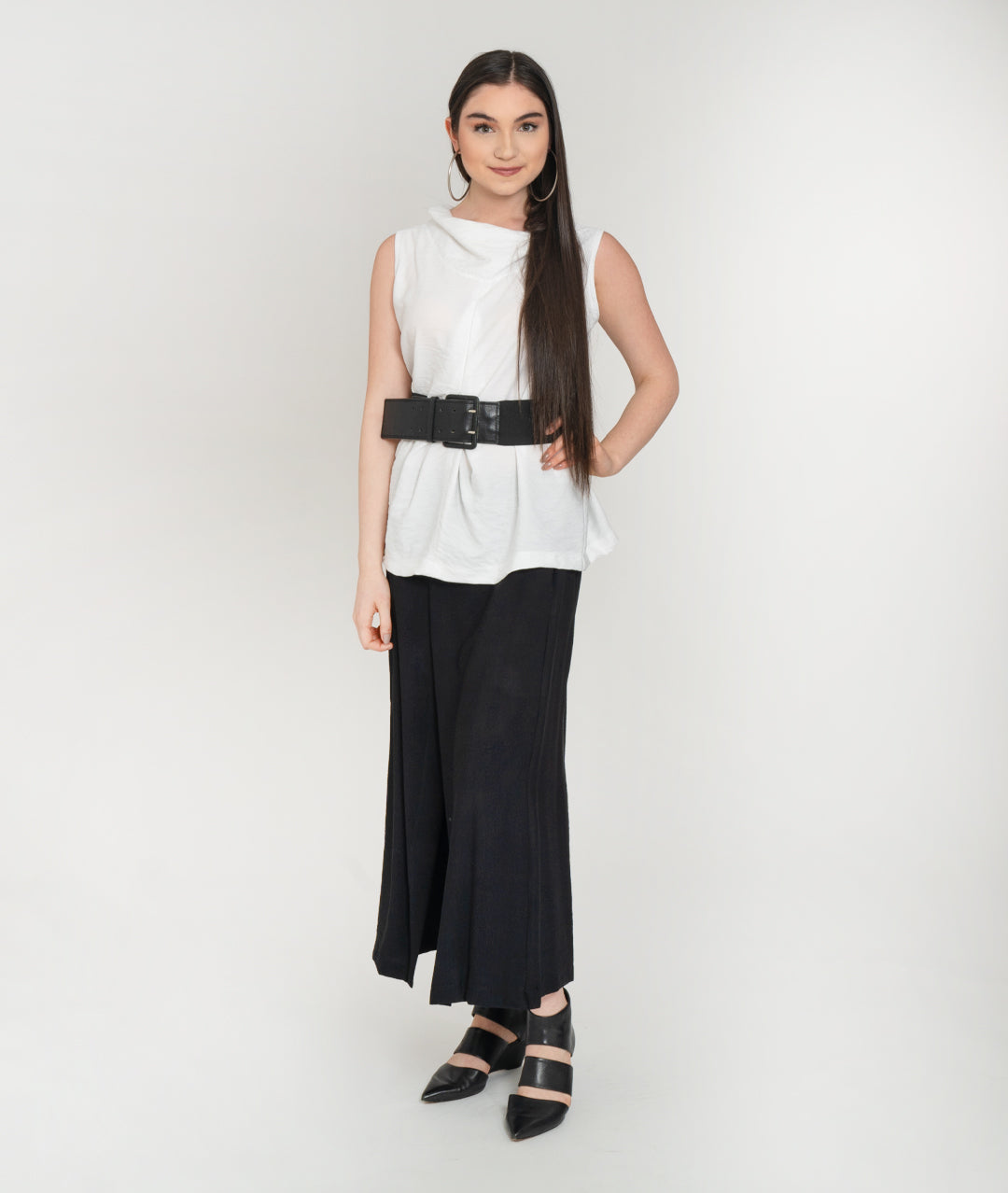 model in a sleeveless white tank with a cowl neck and black belt, worn over a wide leg black pant with an overlapping overlay on either front