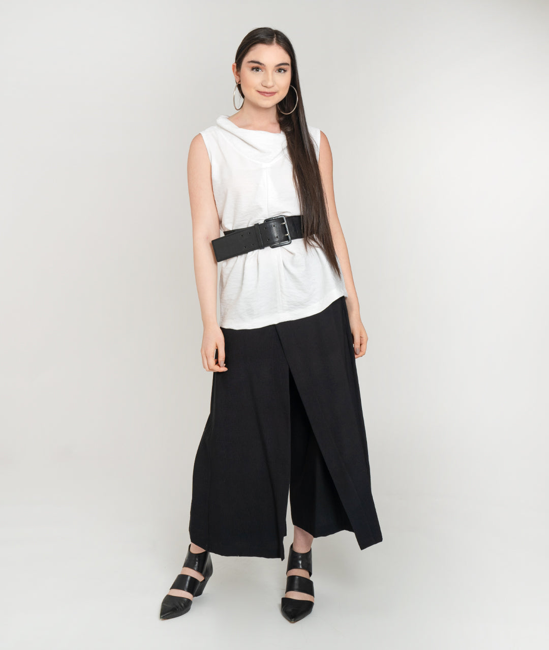 model in a sleeveless white tank with a cowl neck and black belt, worn over a wide leg black pant with an overlapping overlay on either front