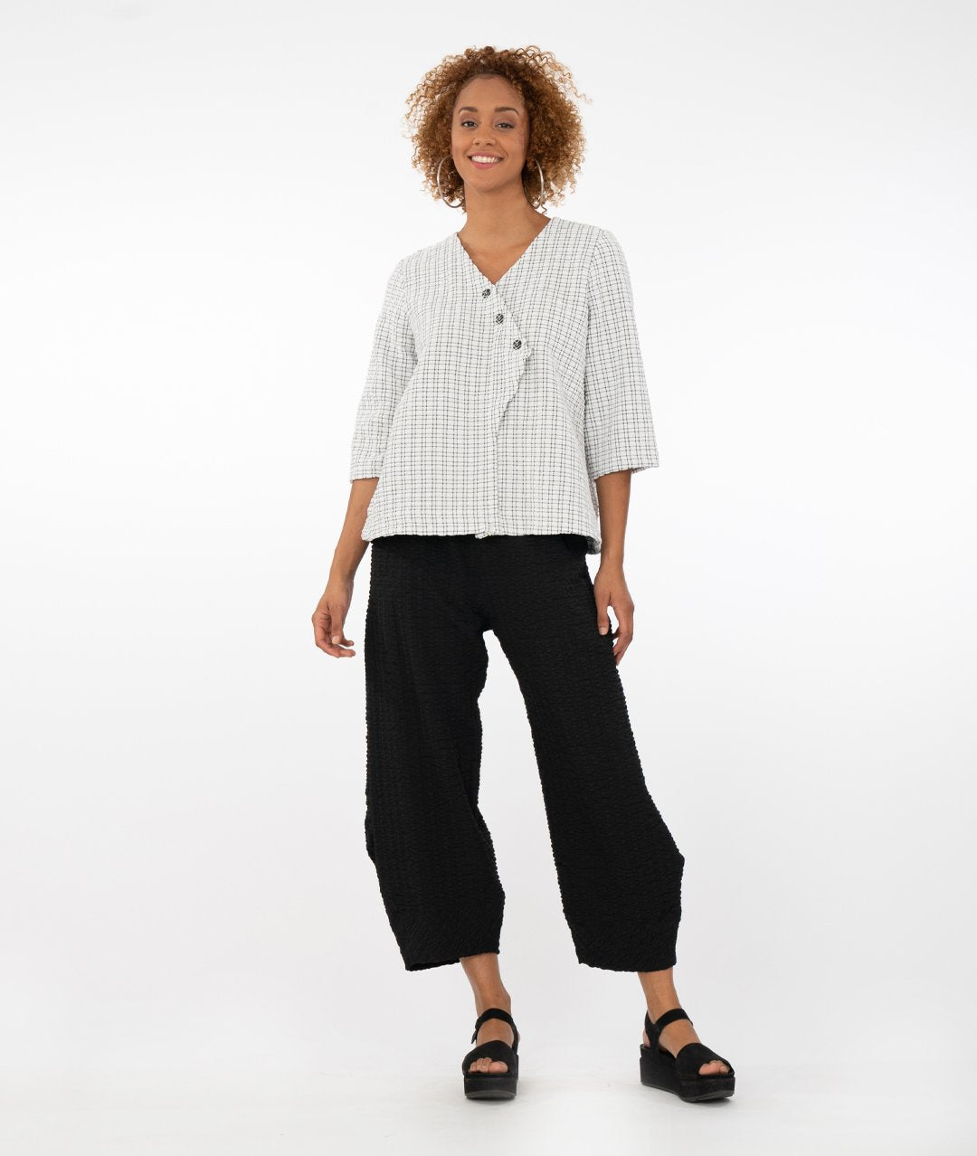 model wearing a black and white checked top with buttons at the front triangular overlap with black pants in front of a white background