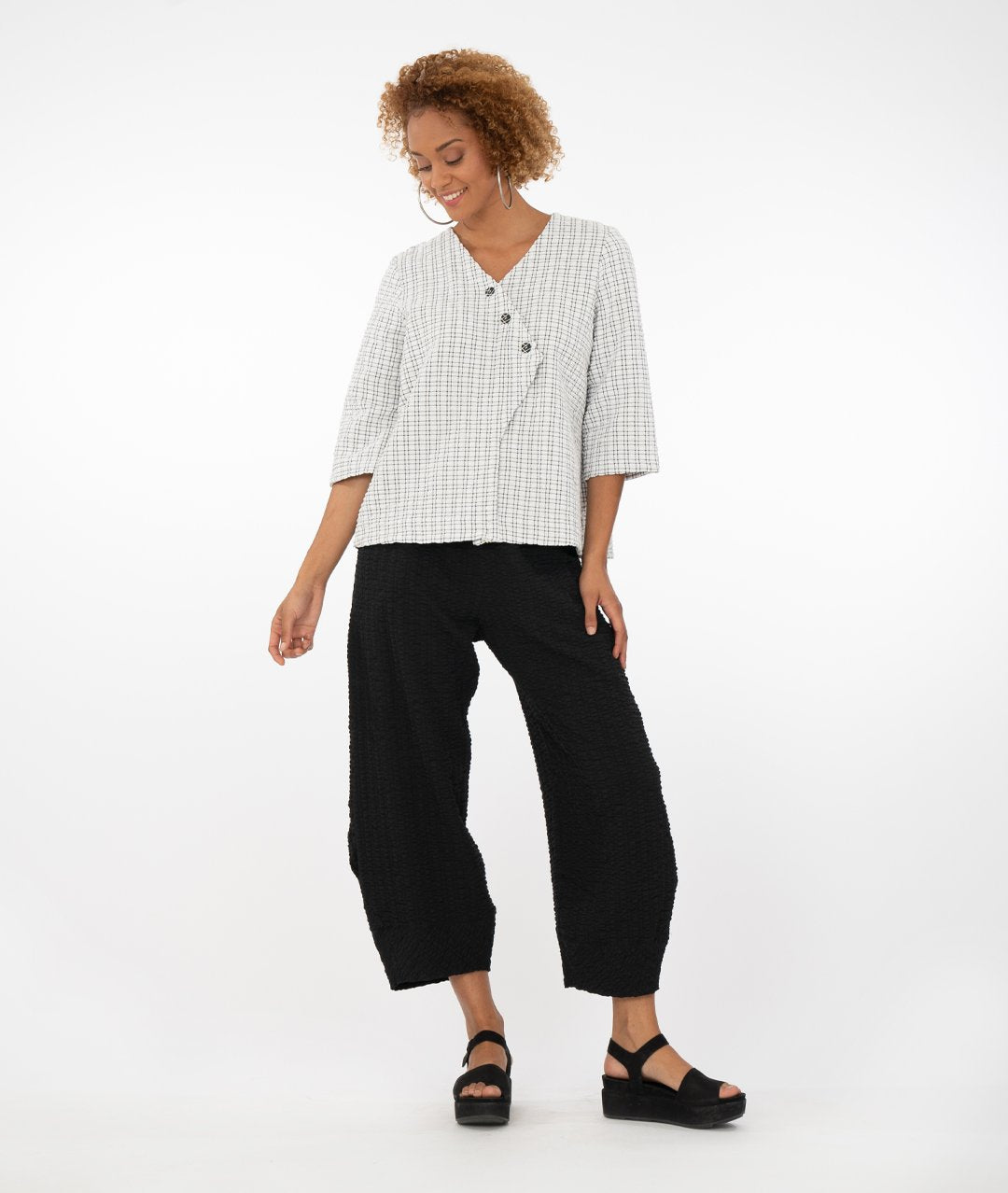 model wearing a black and white checked top with buttons at the front triangular overlap with black pants in front of a white background