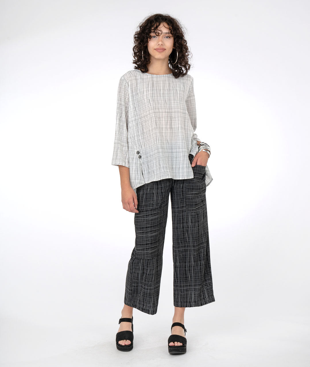 model in a black grid print pant with a white pullover top with a black grid print. top has 3/4 sleeves, godets extending from the sides towards the center front, with a split at each seam topped with a button detail