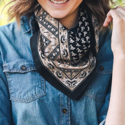Pictured is a photo of a woman smiling and wearing a bandana scarf around her neck. The scarf is black and features tan and white floral vintage motif throughout.