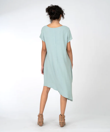 Back view of brunette model wearing light blue assymmterical short sleeve dress with gather at the shoulder. On a white background.