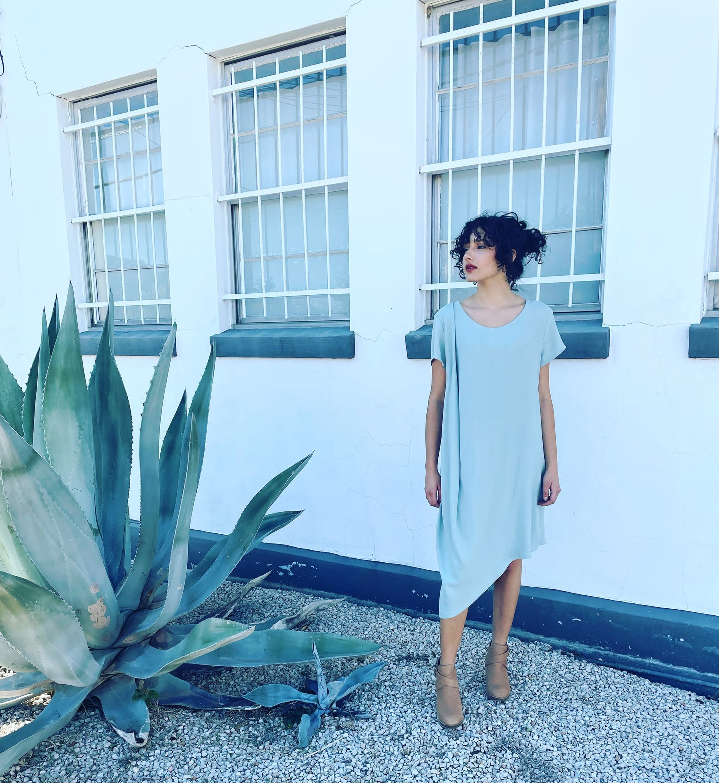 brunette model wearing light blue assymmterical short sleeve dress with gather at the shoulder. On a white building with gravel ground and a large agave plant background.