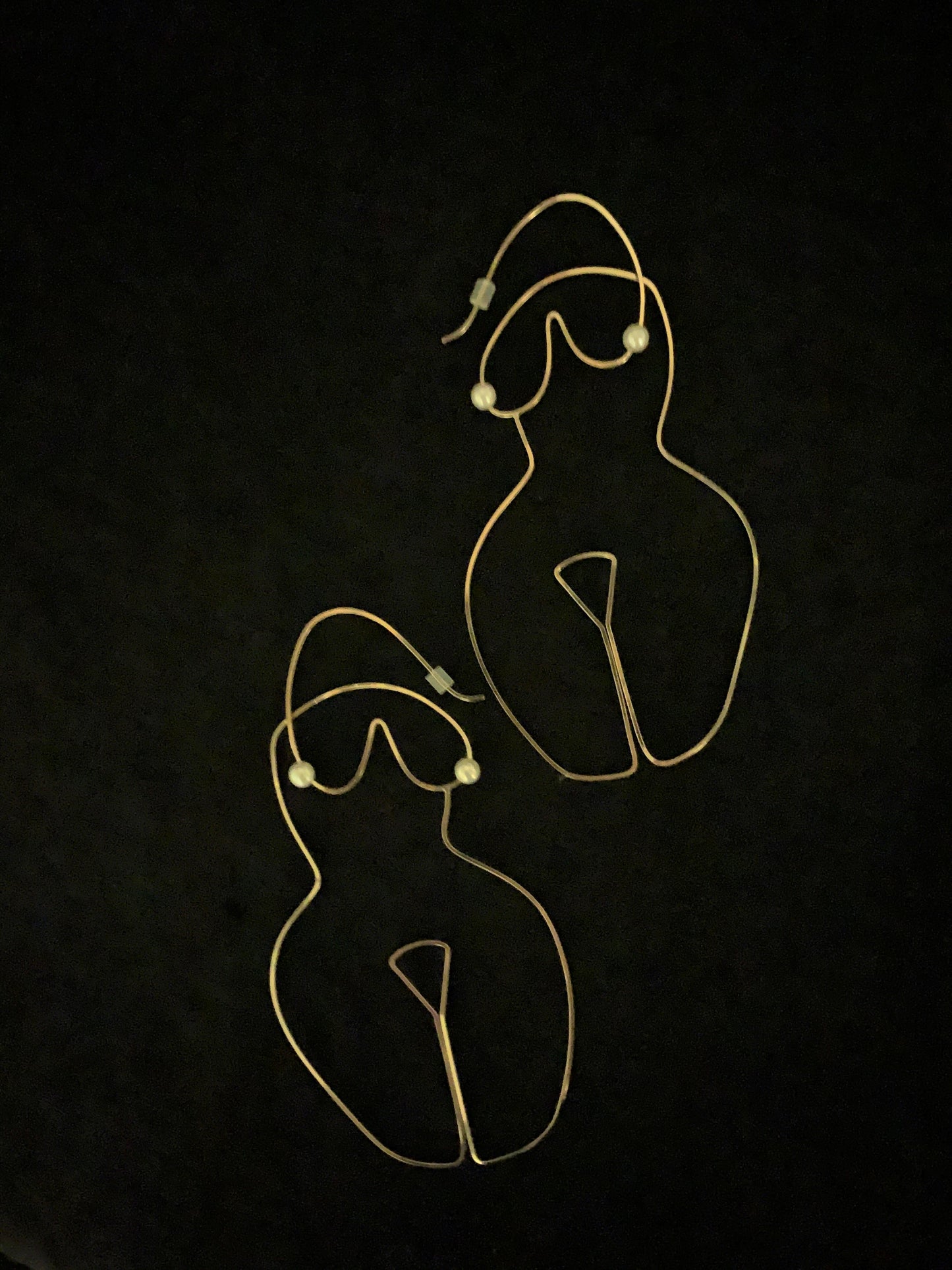 Gold wire earrings shaped in the form of a woman's body with pearl accents as nipples against a black background.