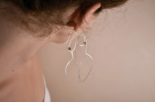 Silver wired earrings in the form a woman's body on the ears of a caucasian model. 