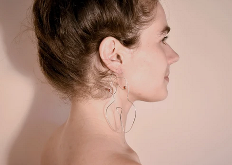 Silver wired earrings in the form a woman's body on the ears of a caucasian model. 