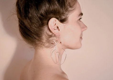 Silver wire earrings shaped into the form of a woman's body on a caucasian model's ears.
