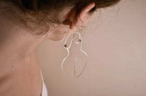 Silver wire earrings shaped into the form of a woman's body on a caucasian model's ears.
