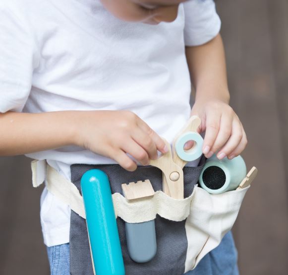 Young child wearing a white top and denim bottoms. He is also wearing the navy blue utility belt with different wooden hair dresser tools in it.