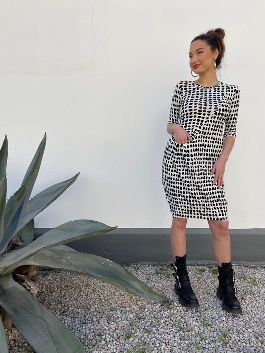 Model in Blck and white polka dot tunic with front pocket and round neckline. Front pocket and hits above the knee. Against a white background, gravel ground and agave plant to the left.