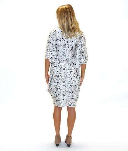 Back side blond model wearing a v-neck kaftan style dress in a white terrazzo print and taupe shoes. Against a White background