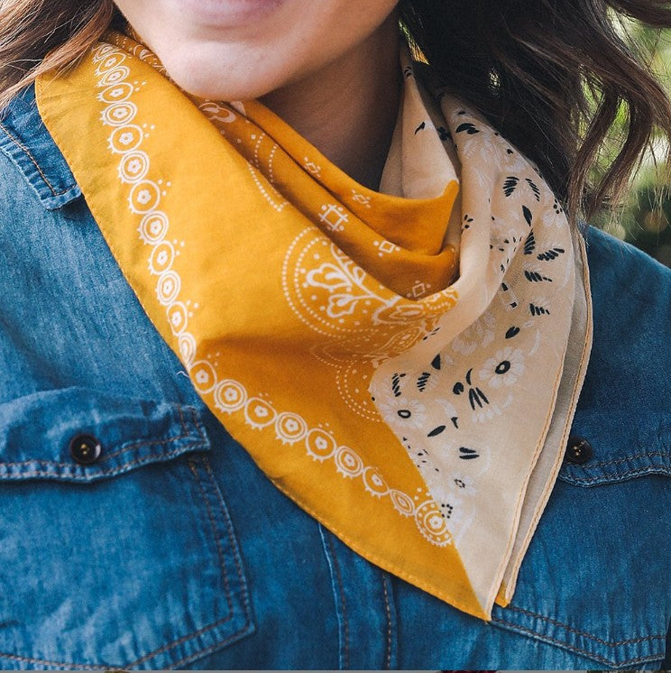 close up of model wearing a yellow and mustard bandana with black floral print tied around her neck. Against a drak denim jacket. Only her chin and ends of her brown hair are visibible in the photo.