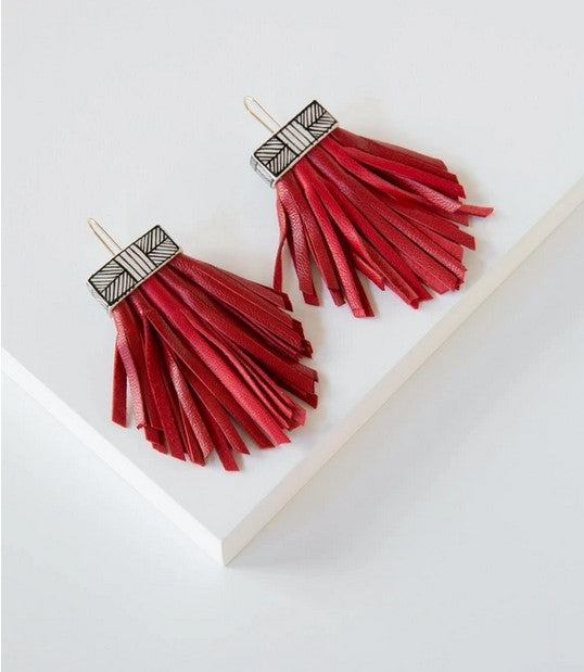 Pictured against a white background are earrings with gold wire hooks, rectangle shaped ceramic bases painted with white and black stripes, and long leather tassels that are hand cut and red leather.